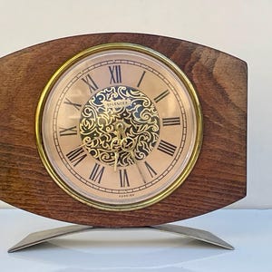 May include: A vintage wooden clock with a gold-tone bezel and a cream-colored face featuring Roman numerals. The clock has an ornate, openwork design in the center and sits on a curved, silver-tone metal base.