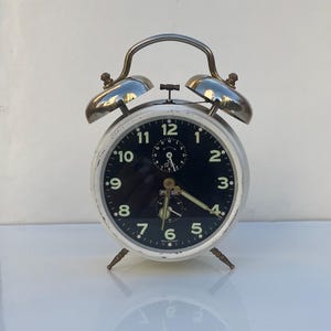 May include: A vintage alarm clock with a black face, white numerals, and a silver metal frame. The clock features two silver bells and small legs. The hands are visible, and the clock face has a small central dial.