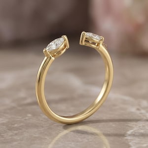 May include: A gold-colored ring with two marquise-cut diamonds set at the ends of the open band. The ring's design features a split shank, creating an open space between the diamonds. The ring is set on a neutral-toned surface.