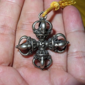 Pode incluir: Um pingente de metal em tom prateado de vajra duplo com um design em forma de cruz e detalhes ornamentais intrincados em cada um dos quatro braços. Um pequeno laço de metal no topo conecta-se a um cordão amarelo brilhante para suspensão.
