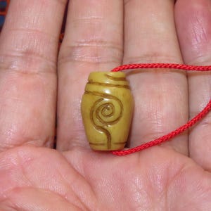 Puede incluir: Un pequeño colgante en forma de barril de color amarillo claro, con un diseño de espiral tallado. El colgante está suspendido de un cordón rojo brillante, creando un contraste llamativo. Probablemente sea una joya o un objeto decorativo.