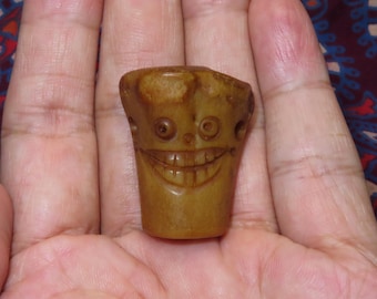 May include: A small, carved wooden figure with a smiling face. The figure is a light brown color and has a cylindrical shape. The face features carved eyes, a nose, and a wide, toothy grin. The figure is held in a person's hand.