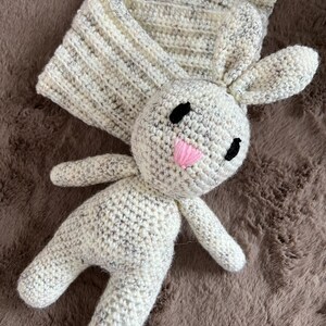 May include: A crocheted white bunny with black eyes and a pink nose. The bunny is lying on a brown fuzzy surface.