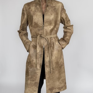 May include: A tan-coloured coat with a textured surface and a zip-up front. The coat features a high collar, long sleeves, and a matching belt. The coat is knee-length and has two front pockets.