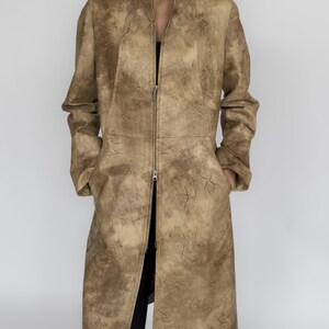 May include: A long, tan coat with a high collar and a full-length zip. The coat has a textured, crackled pattern throughout. The sleeves are long, and the coat reaches to the mid-calf. The coat is worn over a black top and trousers.