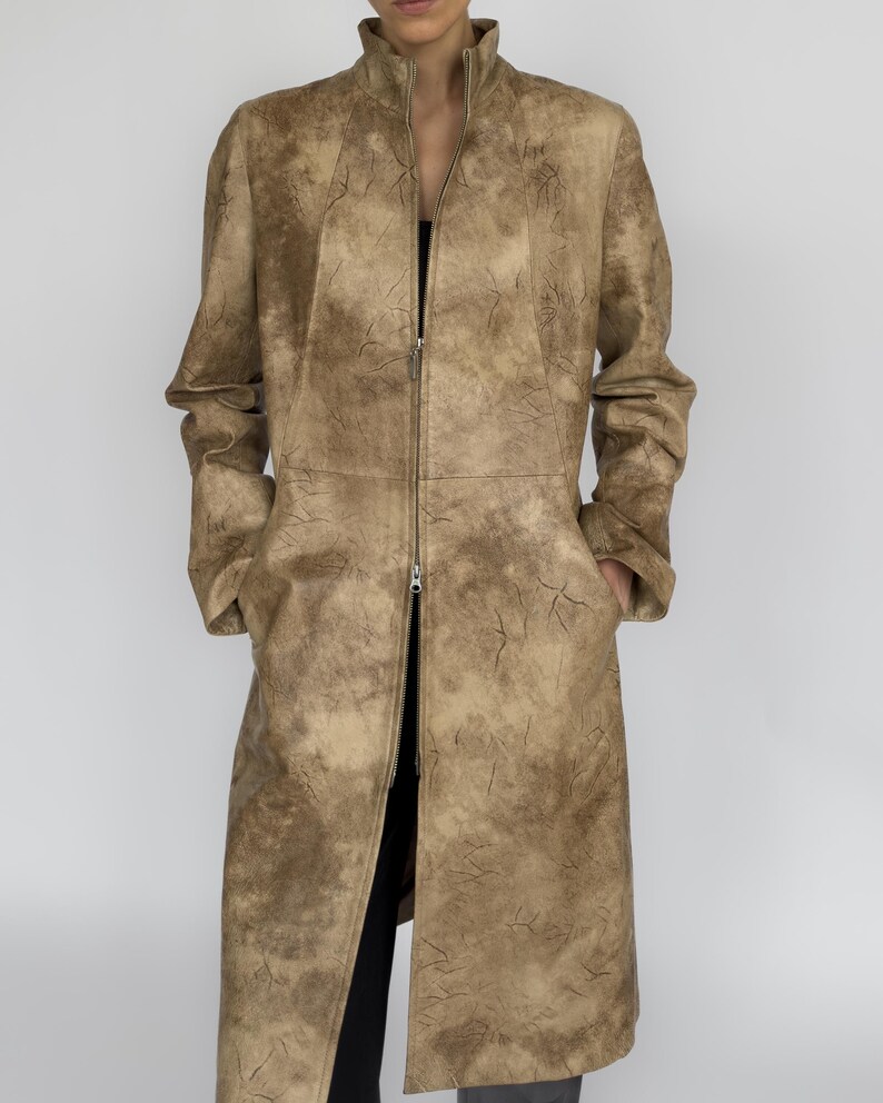 May include: A long, tan coat with a high collar and a full-length zip. The coat has a textured, crackled pattern throughout. The sleeves are long, and the coat reaches to the mid-calf. The coat is worn over a black top and trousers.