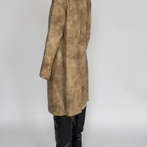 May include: A long, tan coat with a textured, mottled pattern. The coat has a high collar and long sleeves. The person is wearing black boots. The coat appears to be made of leather or a similar material.