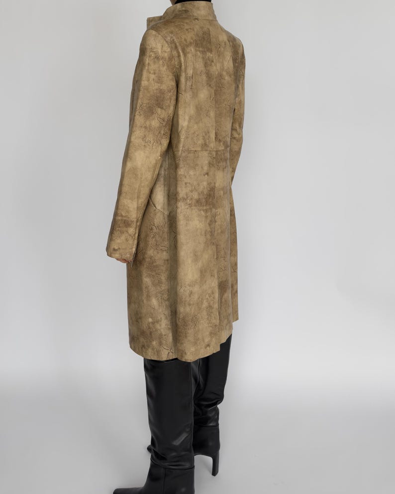 May include: A long, tan coat with a textured, mottled pattern. The coat has a high collar and long sleeves. The person is wearing black boots. The coat appears to be made of leather or a similar material.