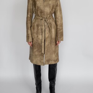May include: A long, tan leather coat with a zip-up front and a matching belt. The coat has a textured surface and is worn with black knee-high boots. The coat is knee-length and has long sleeves.