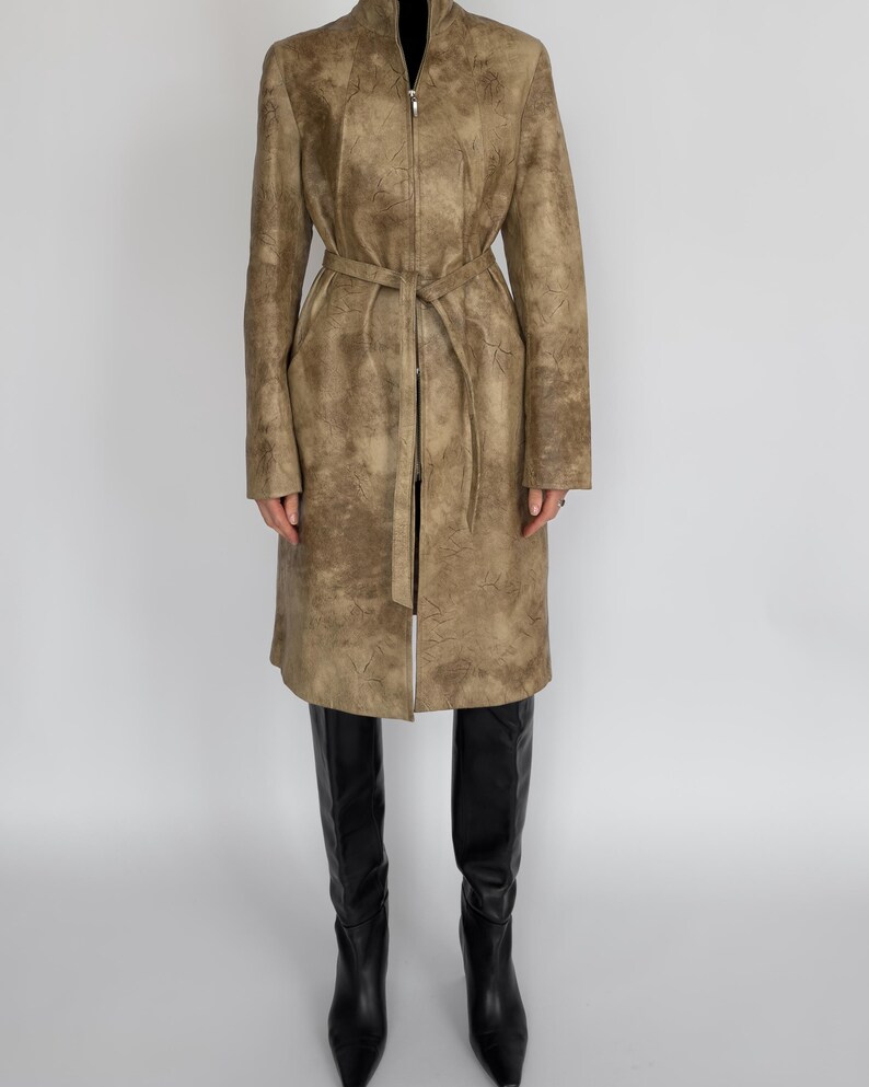 May include: A long, tan leather coat with a zip-up front and a matching belt. The coat has a textured surface and is worn with black knee-high boots. The coat is knee-length and has long sleeves.