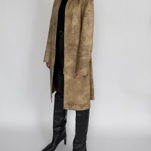 May include: A long, tan coat with a textured pattern, worn over a black turtleneck and black trousers. The coat has a high collar and a full-length zip. Black knee-high boots complete the outfit.