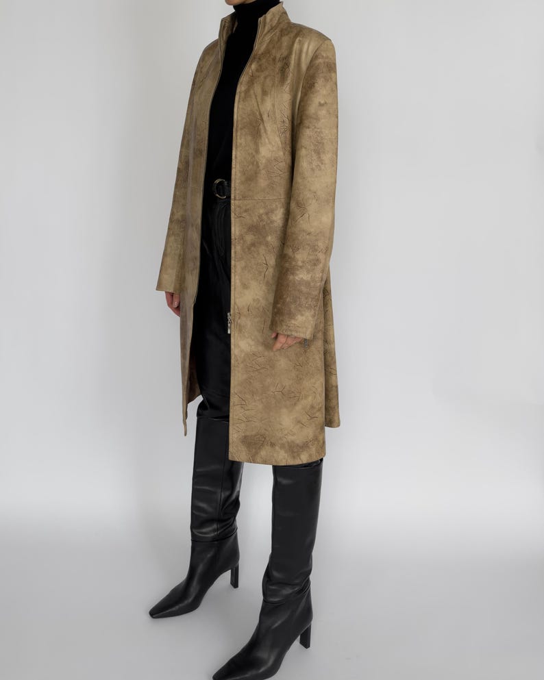 May include: A long, tan coat with a textured pattern, worn over a black turtleneck and black trousers. The coat has a high collar and a full-length zip. Black knee-high boots complete the outfit.