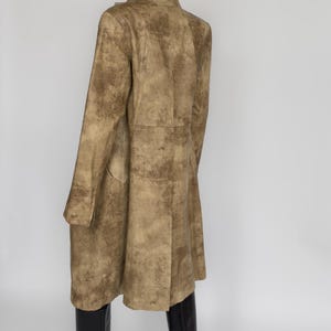 May include: A long, tan coat with a textured pattern. The coat has a button closure and long sleeves. The model is wearing black boots. The coat appears to be made of a soft material.