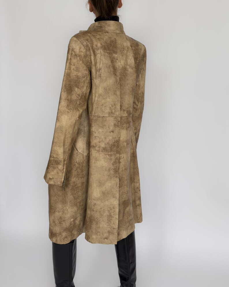 May include: A long, tan coat with a textured pattern. The coat has a button closure and long sleeves. The model is wearing black boots. The coat appears to be made of a soft material.