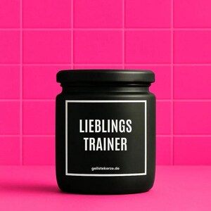 Op de afbeelding: Een matzwarte cilindrische kaarsenpot met een bijpassend zwart deksel. De voorzijde is voorzien van een witte vierkante rand met de tekst LIEBLINGS TRAINER in dikke witte hoofdletters, met de website geilstekerze.de in kleine witte letters eronder.
