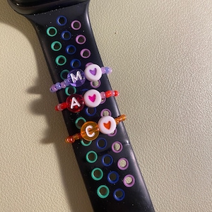 Puede incluir: Correa de reloj inteligente negra con tres anillos de cuentas. Cada anillo presenta una cuenta de letra y una cuenta de corazón. Las letras son M, A y C. Las cuentas son en tonos morado, rojo y naranja. La esfera del reloj muestra un teclado numérico digital.