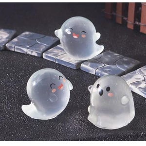 Pode incluir: Tr&ecirc;s figuras transl&uacute;cidas de fantasmas com rostos sorridentes e bochechas rosadas. Os fantasmas est&atilde;o em poses diferentes, com um flutuando sobre uma telha de pedra. O fundo apresenta mais telhas de pedra e uma superf&iacute;cie escura.
