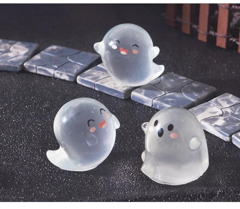 Pode incluir: Tr&ecirc;s figuras transl&uacute;cidas de fantasmas com rostos sorridentes e bochechas rosadas. Os fantasmas est&atilde;o em poses diferentes, com um flutuando sobre uma telha de pedra. O fundo apresenta mais telhas de pedra e uma superf&iacute;cie escura.