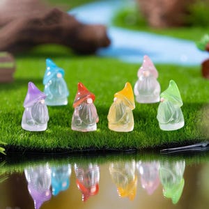 Puede incluir: Una colección de gnomos de jardín en miniatura con cuerpos translúcidos y sombreros coloridos. Los gnomos están dispuestos sobre un lecho de hierba verde, con reflejos visibles debajo. Los sombreros son de varios colores, incluyendo morado, azul, rojo, amarillo, rosa y verde.