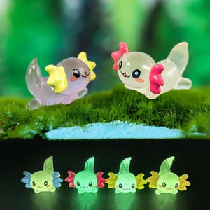 Puede incluir: Una colección de pequeñas figuras translúcidas de ajolotes en varios colores. Las figuras tienen caras bonitas y de dibujos animados y están posadas sobre un lecho de musgo verde. Algunas tienen detalles rosas, amarillos y azules.