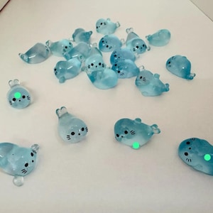 May include: A scattering of light blue seal figurines with black eyes and whiskers. The seals have a translucent appearance and are arranged on a white surface. Some seals have a green dot on their belly. These miniature seals are likely decorative items.
