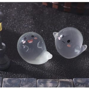 Pode incluir: Duas figuras de fantasmas cinzentos transl&uacute;cidos com bochechas rosadas e tra&ccedil;os faciais pretos. Um fantasma tem um sorriso de boca aberta, o outro um sorriso de boca fechada. Os fantasmas est&atilde;o numa superf&iacute;cie escura.