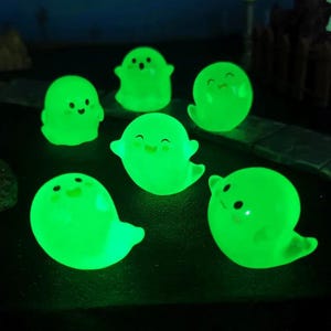 Pode incluir: Uma cole&ccedil;&atilde;o de sete figuras de fantasmas verdes brilhantes com rostos sorridentes. As figuras t&ecirc;m uma forma arredondada com pequenos bra&ccedil;os e uma base em forma de cauda. Os fantasmas est&atilde;o dispostos sobre uma superf&iacute;cie escura.