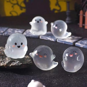 Pode incluir: Uma cole&ccedil;&atilde;o de seis figuras de fantasmas transl&uacute;cidos com rostos sorridentes e bochechas rosadas. Os fantasmas t&ecirc;m uma forma arredondada com bra&ccedil;os pequenos e uma parte inferior fluida. Est&atilde;o dispostos sobre uma superf&iacute;cie escura com ladrilhos de pedra no fundo.