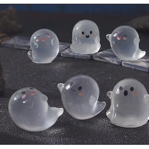 Pode incluir: Seis figuras transl&uacute;cidas de fantasmas com rostos sorridentes e bochechas rosadas. Os fantasmas t&ecirc;m bra&ccedil;os pequenos e uma parte inferior fluida. Est&atilde;o dispostos sobre uma superf&iacute;cie escura, sugerindo um tema de Halloween ou assustador.