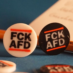 Könnte beinhalten: Eine Ansammlung weißer und schwarzer runder Objekte mit dem Text "FCK AFD" in fetter Schrift, durchgestrichen mit einem roten Strich. Die Objekte liegen auf einer blauen Oberfläche, im Hintergrund Geld und ein Bleistift.