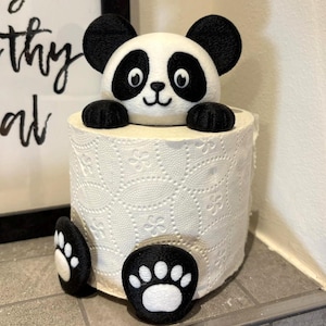 Könnte beinhalten: Eine weiße Toilettenpapierrolle mit floralem Muster, verziert mit einem Plüsch-Pandabärenkopf und -pfoten. Der Panda hat schwarze Ohren, Augen und Pfoten und ein weißes Gesicht. Der Panda umarmt die Toilettenpapierrolle.