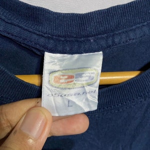 K&ouml;nnte beinhalten: Ein marineblaues T-Shirt mit einem wei&szlig;en Etikett, auf dem "B.U.S.A. Inc." und "L" steht.