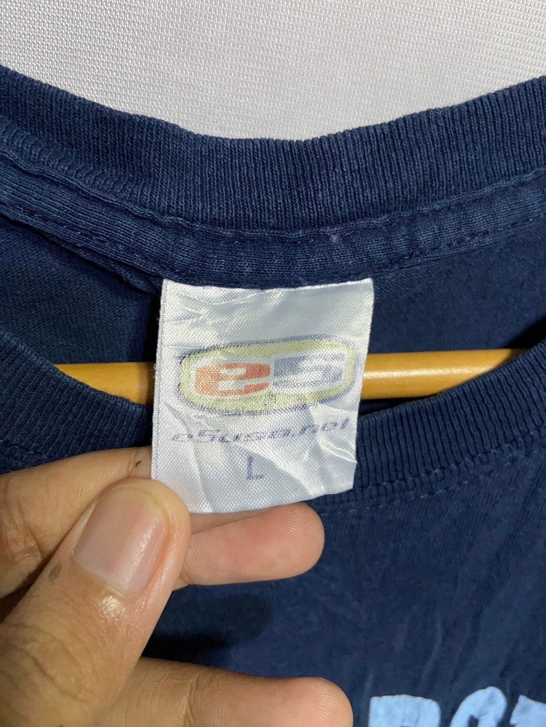 K&ouml;nnte beinhalten: Ein marineblaues T-Shirt mit einem wei&szlig;en Etikett, auf dem "B.U.S.A. Inc." und "L" steht.