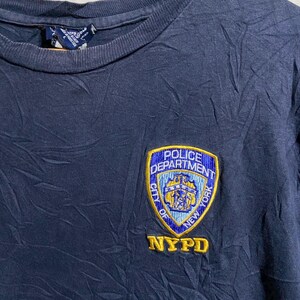 Peut inclure: T-shirt bleu marine avec un écusson brodé du NYPD Police Department. L'écusson est bleu, jaune et blanc, avec les mots "Police Department City of New York" et "NYPD" en or.