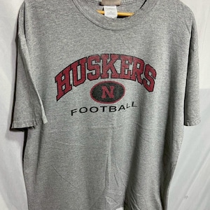 Op de afbeelding: Een grijs t-shirt met het woord "Huskers" in rode blokletters boven een rood en zwart ovaal met de letter "N". Onder het ovaal staat het woord "FOOTBALL" in het zwart gedrukt. Het shirt hangt aan een houten hanger.