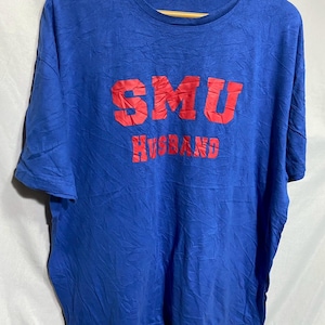 May include: A blue t-shirt with the red text "SMU HUSBAND" printed on the front. The shirt is hanging on a wooden hanger. The fabric has a textured appearance.