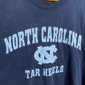 K&ouml;nnte beinhalten: Ein marineblaues T-Shirt mit einem hellblauen Aufdruck des Logos der North Carolina Tar Heels und dem Text "NORTH CAROLINA TAR HEELS."