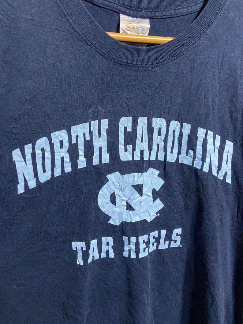 K&ouml;nnte beinhalten: Ein marineblaues T-Shirt mit einem hellblauen Aufdruck des Logos der North Carolina Tar Heels und dem Text "NORTH CAROLINA TAR HEELS."