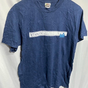 Può includere: T-shirt blu navy a maniche corte con la scritta "TRINITY CHRISTIAN COLLEGE" in bianco e blu. La maglietta è appesa a una gruccia di legno. La maglietta è leggermente stropicciata.