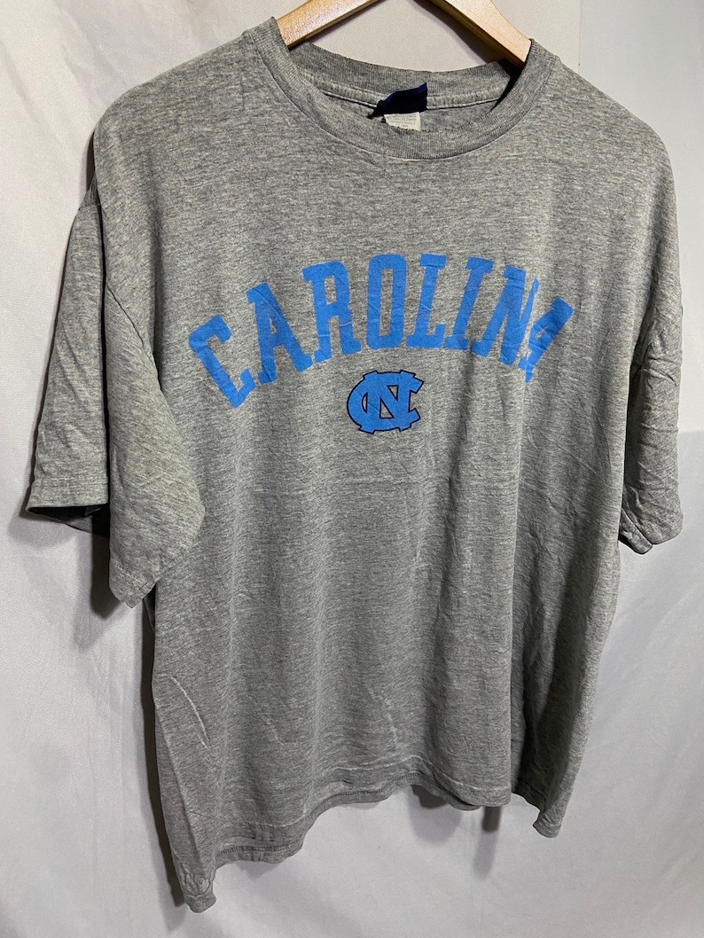 Puede incluir: Camiseta gris con un dise&ntilde;o de arco azul "CAROLINA" y un logotipo azul "UNC".