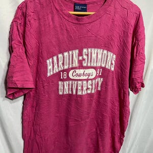 Puede incluir: Camiseta rosa con el texto blanco "HARDIN-SIMMONS UNIVERSITY". Debajo, las palabras "18 Cowboys 91". La camiseta tiene mangas cortas y cuello redondo. La camiseta está colgada en una percha de madera.
