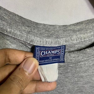 Puede incluir: Camiseta gris con un estampado gr&aacute;fico azul y una etiqueta que dice "Oficialmente Hecho Exclusivamente para Champs Sports con licencia de Team Edition Apparel".