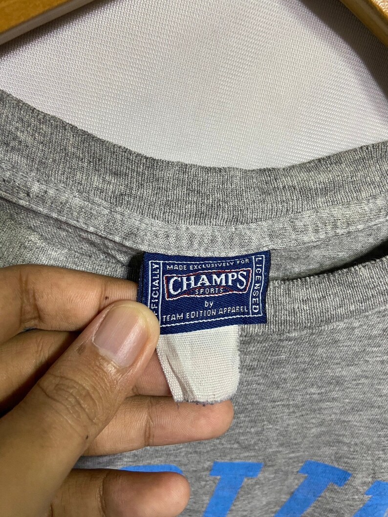 Puede incluir: Camiseta gris con un estampado gr&aacute;fico azul y una etiqueta que dice "Oficialmente Hecho Exclusivamente para Champs Sports con licencia de Team Edition Apparel".