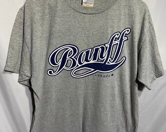Könnte beinhalten: Graues T-Shirt mit einer blauen und weißen Grafik des Wortes "Banff" in einer stilisierten Schriftart. Die Grafik enthält auch das Wort "Kanada" und ein Ahornblatt.