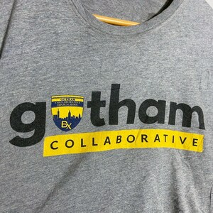 May include: Gray t-shirt with the word "gotham" in large black letters. Below the word "gotham" is the word "COLLABORATIVE" in yellow letters. A small emblem with a cityscape is above the word "gotham".