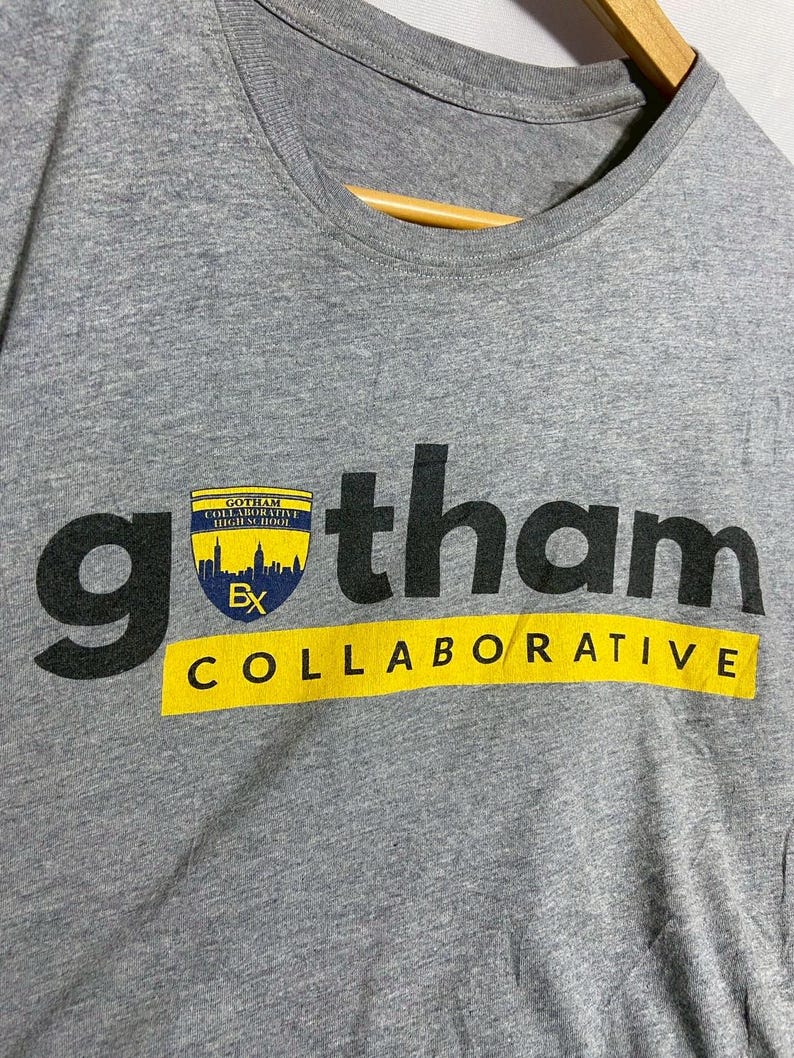 May include: Gray t-shirt with the word "gotham" in large black letters. Below the word "gotham" is the word "COLLABORATIVE" in yellow letters. A small emblem with a cityscape is above the word "gotham".