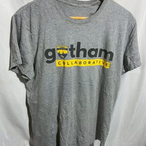 May include: A gray t-shirt with the word "gotham" in large black letters and "COLLABORATIVE" in yellow. A small emblem is above the word "gotham". The shirt is hanging on a wooden hanger.