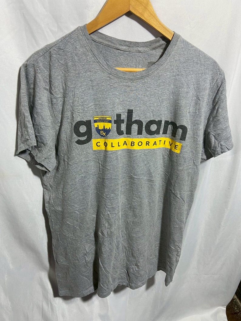 May include: A gray t-shirt with the word "gotham" in large black letters and "COLLABORATIVE" in yellow. A small emblem is above the word "gotham". The shirt is hanging on a wooden hanger.