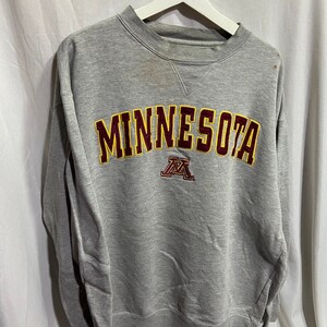 以下が含まれることがあります： グレーのトレーナーに、マロンとイエローの文字で「MINNESOTA」と書いてあり、その下にマロンとイエローの「M」のロゴがあります。