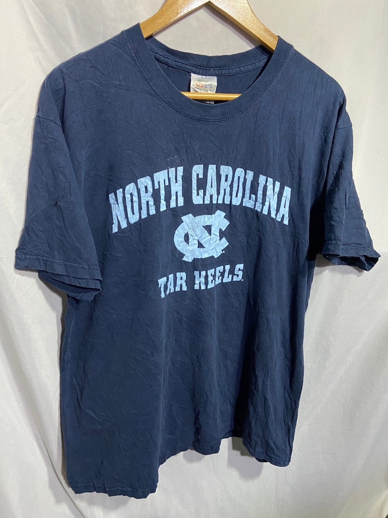 K&ouml;nnte beinhalten: Ein marineblaues T-Shirt mit einem hellblauen Aufdruck des Logos der University of North Carolina Tar Heels und dem Text "NORTH CAROLINA TAR HEELS".
